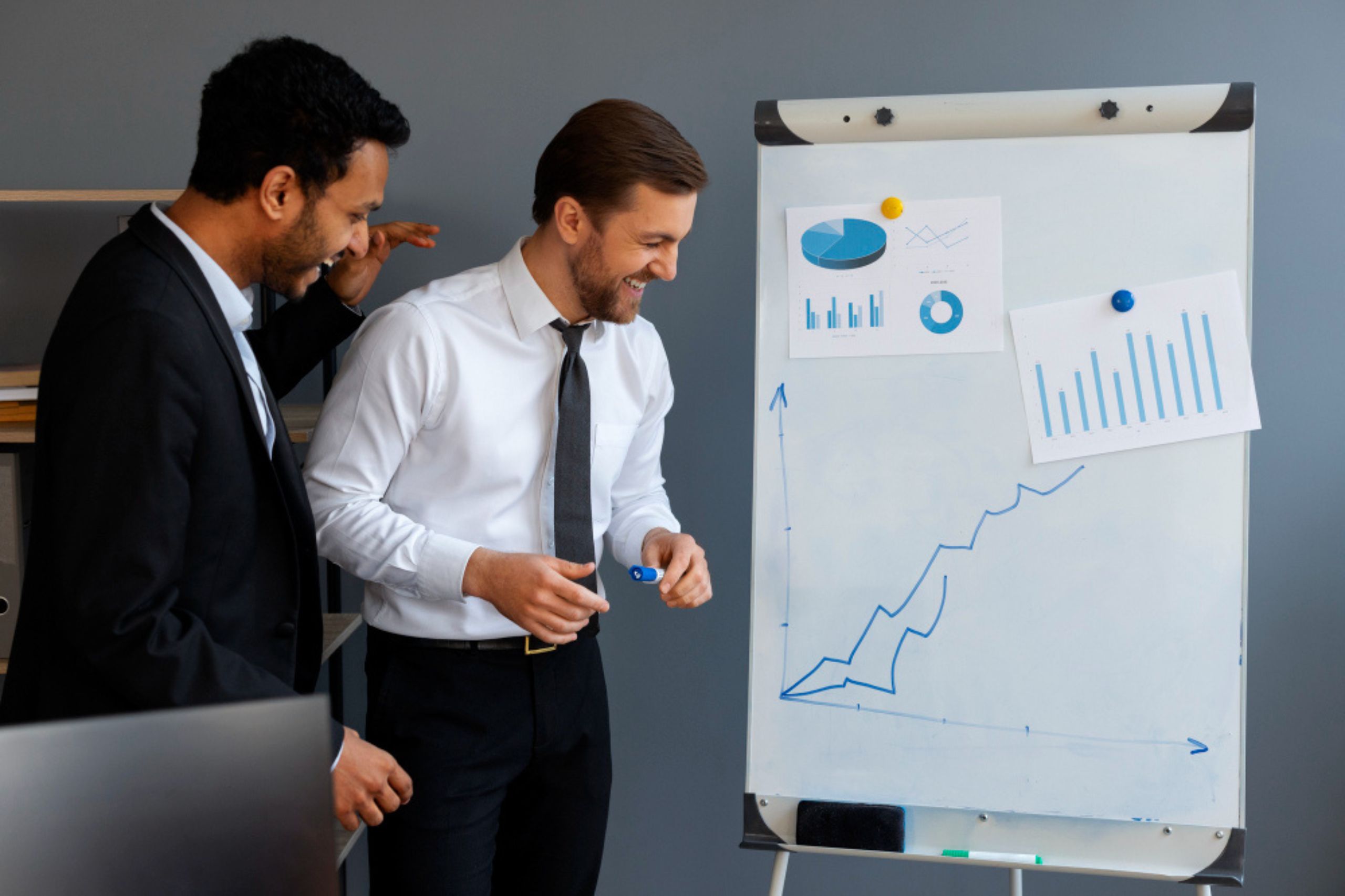 Two men in business attire smile while discussing data and a rising line graph drawn on a flipchart in an office setting.