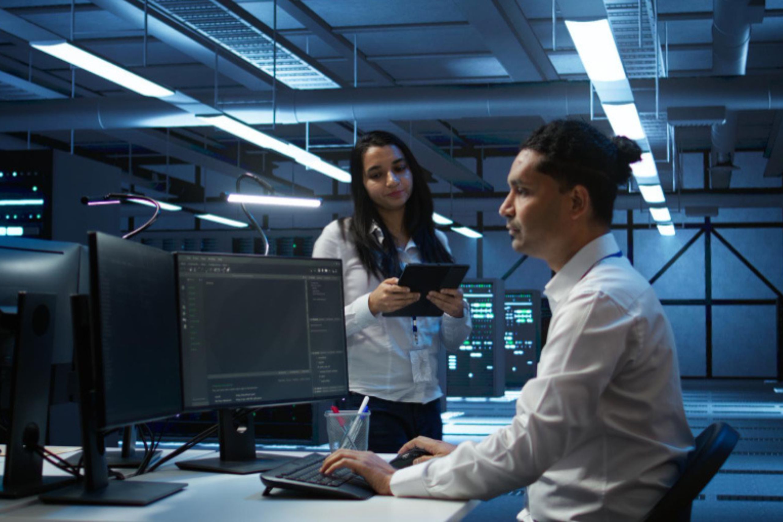 Two IT professionals work in a server room; one sits at a desk with monitors, while the other stands nearby holding a tablet.