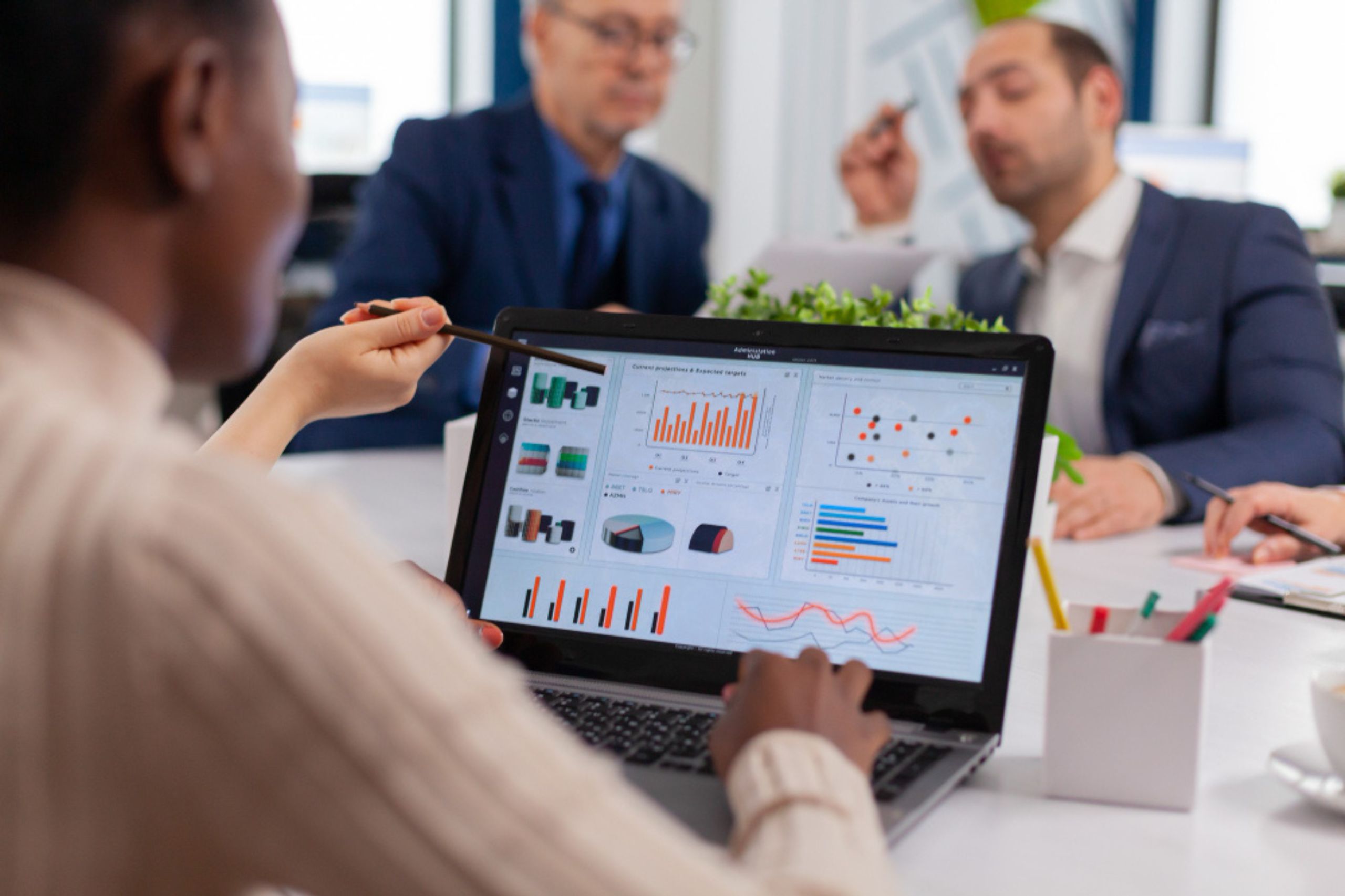 Person holding a pen and analyzing business charts and graphs on a laptop screen during a meeting with two colleagues in an office setting.