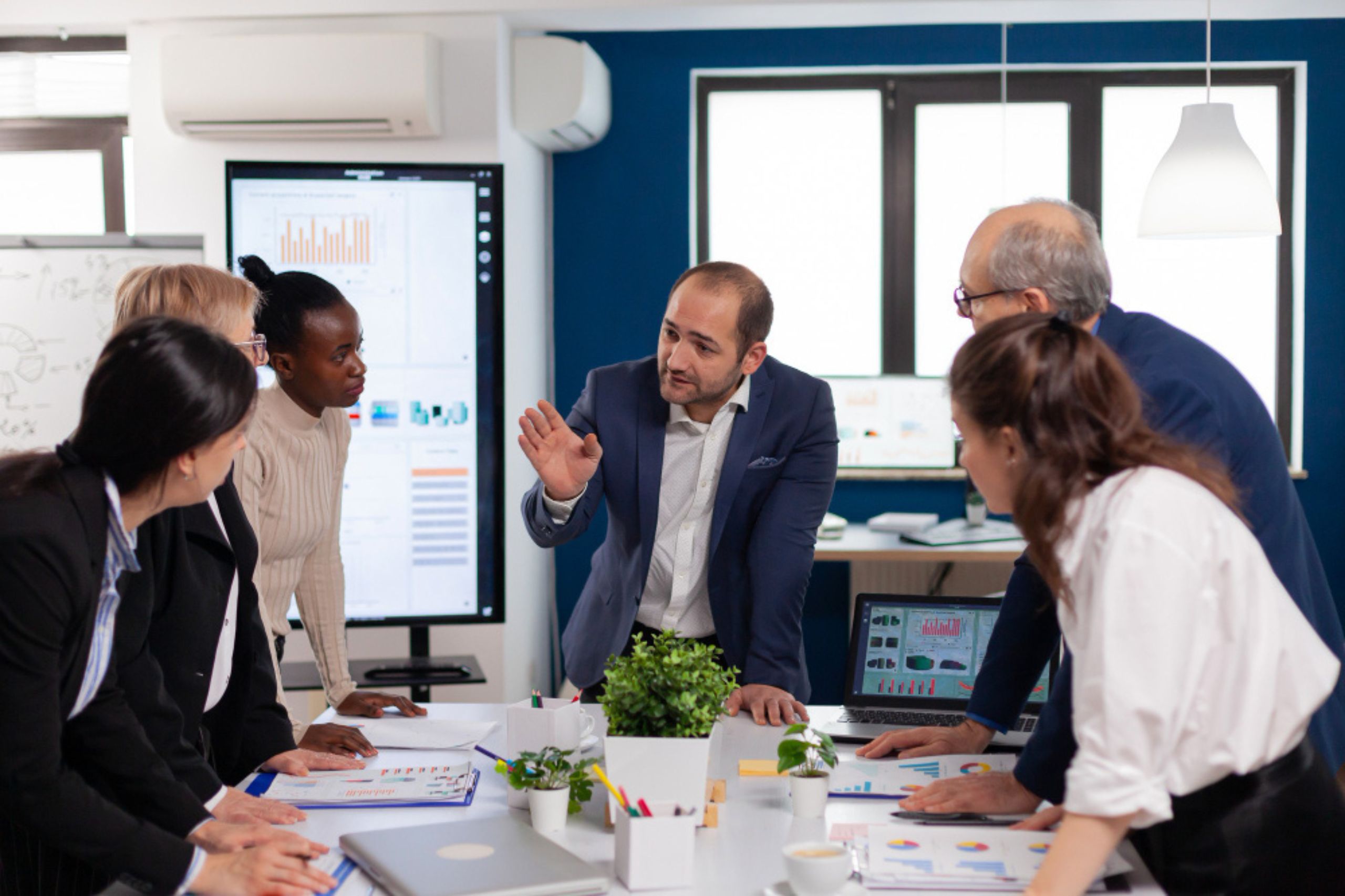 A group of business professionals stand around a conference table, engaged in discussion, with charts and graphs displayed on screens in the background.