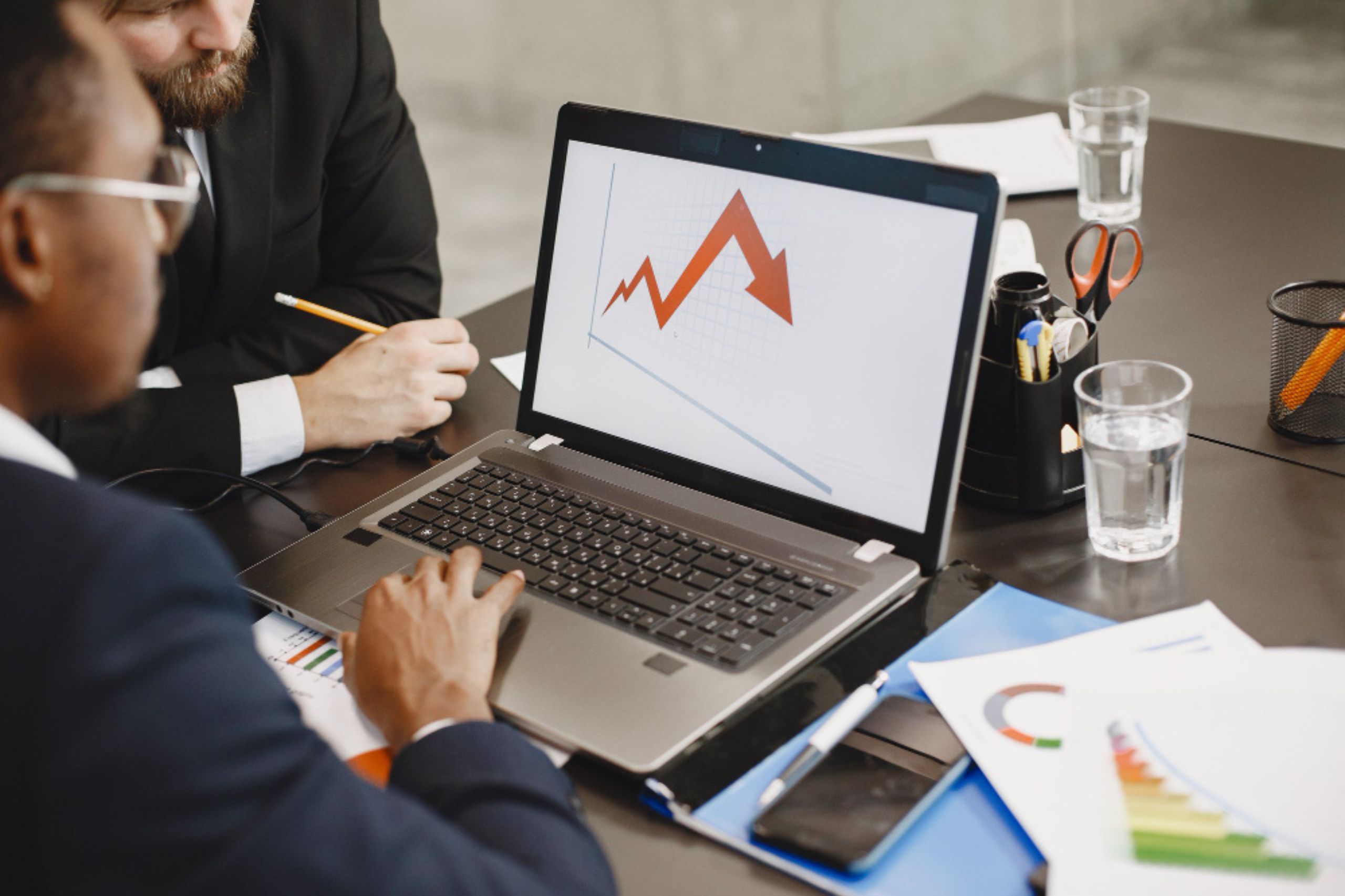 Two people in business attire review a laptop displaying a fluctuating red line graph. Office supplies, documents, and glasses of water are on the black desk.