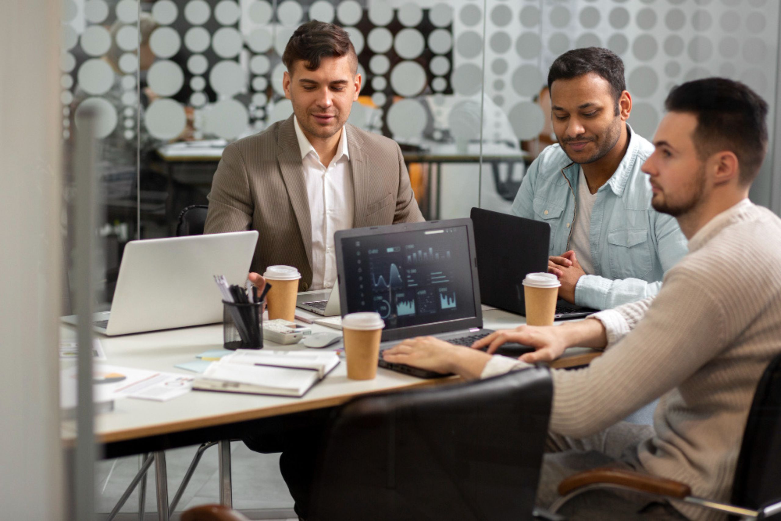 Three men sit at a table in a modern office, working on laptops with charts on the screens and coffee cups beside them.