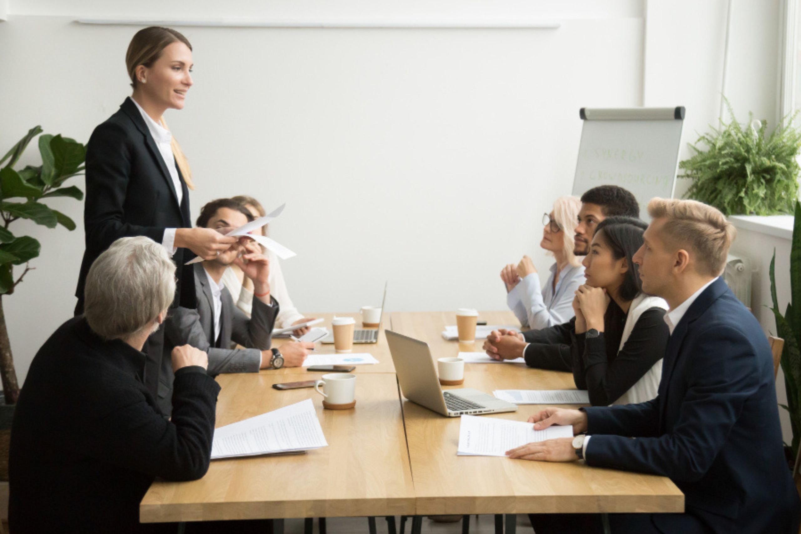 A woman stands and presents to five seated colleagues in a conference room with laptops, papers, and coffee on the table.