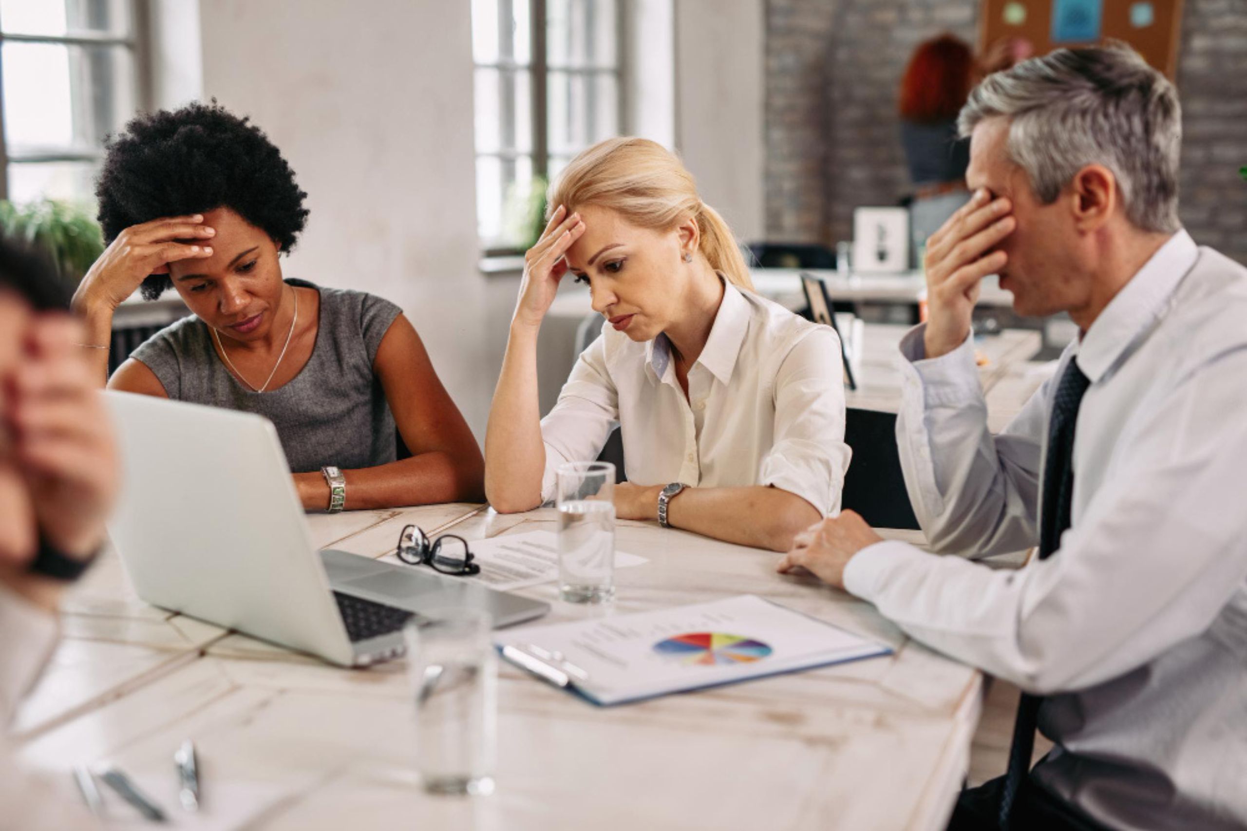Three people sit at a table with a laptop and papers, looking stressed and holding their heads in their hands during a meeting.