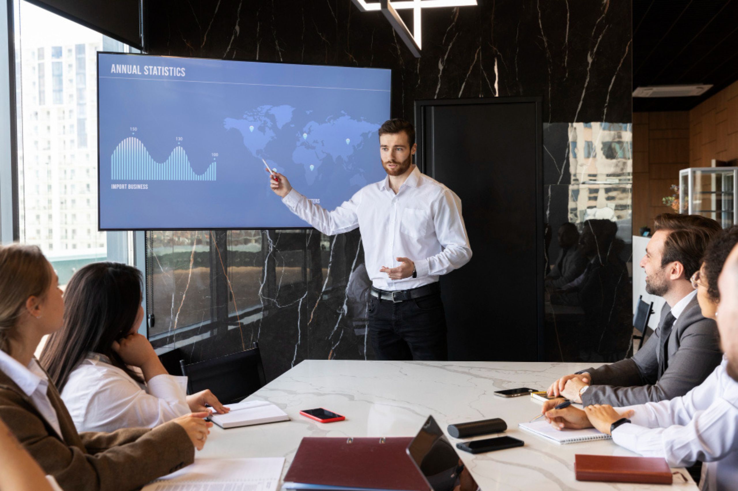 A man in a white shirt presents annual statistics on a screen to four colleagues seated at a conference table in a modern office.