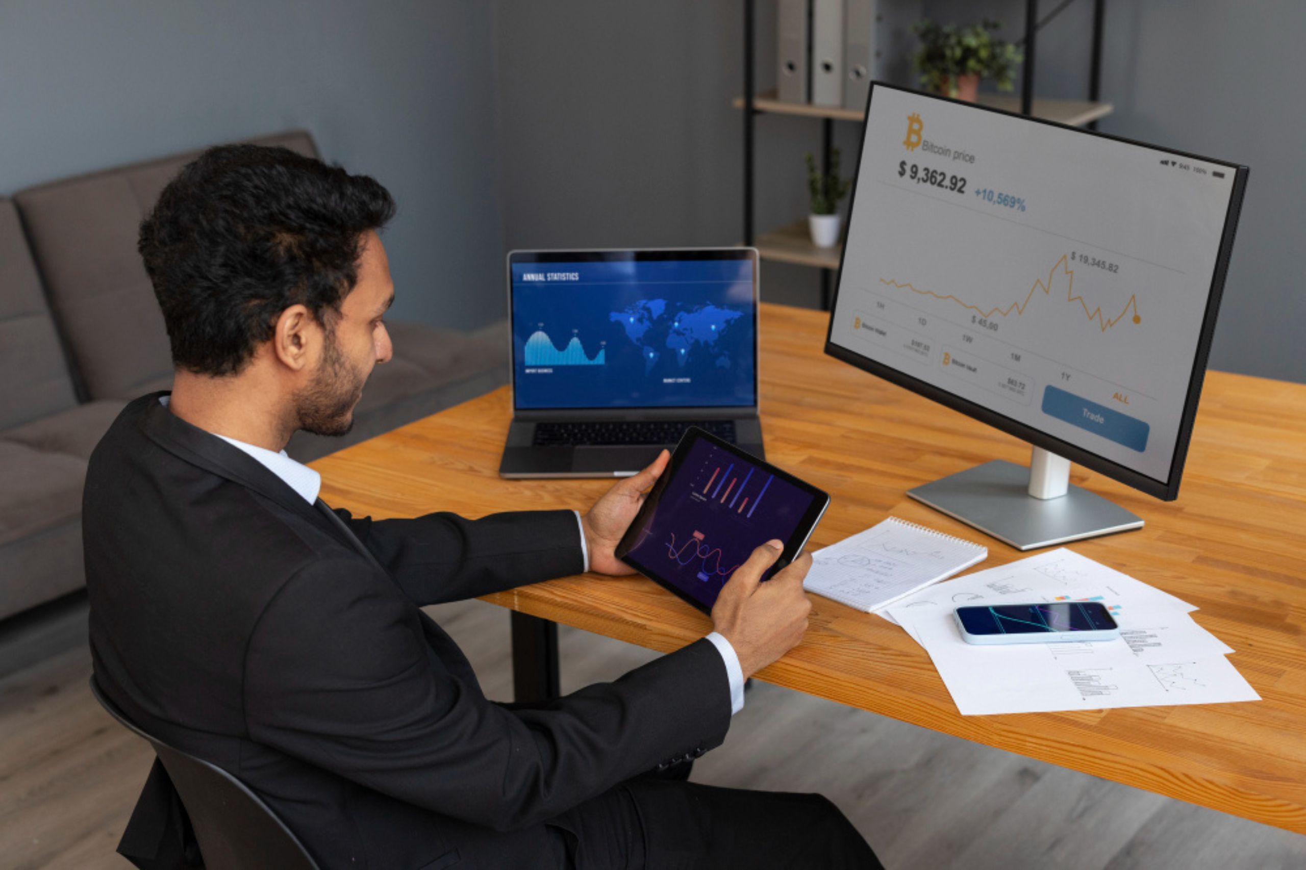 A man in a suit analyzes financial charts on a tablet, with a laptop and desktop monitor displaying cryptocurrency data on a wooden desk.