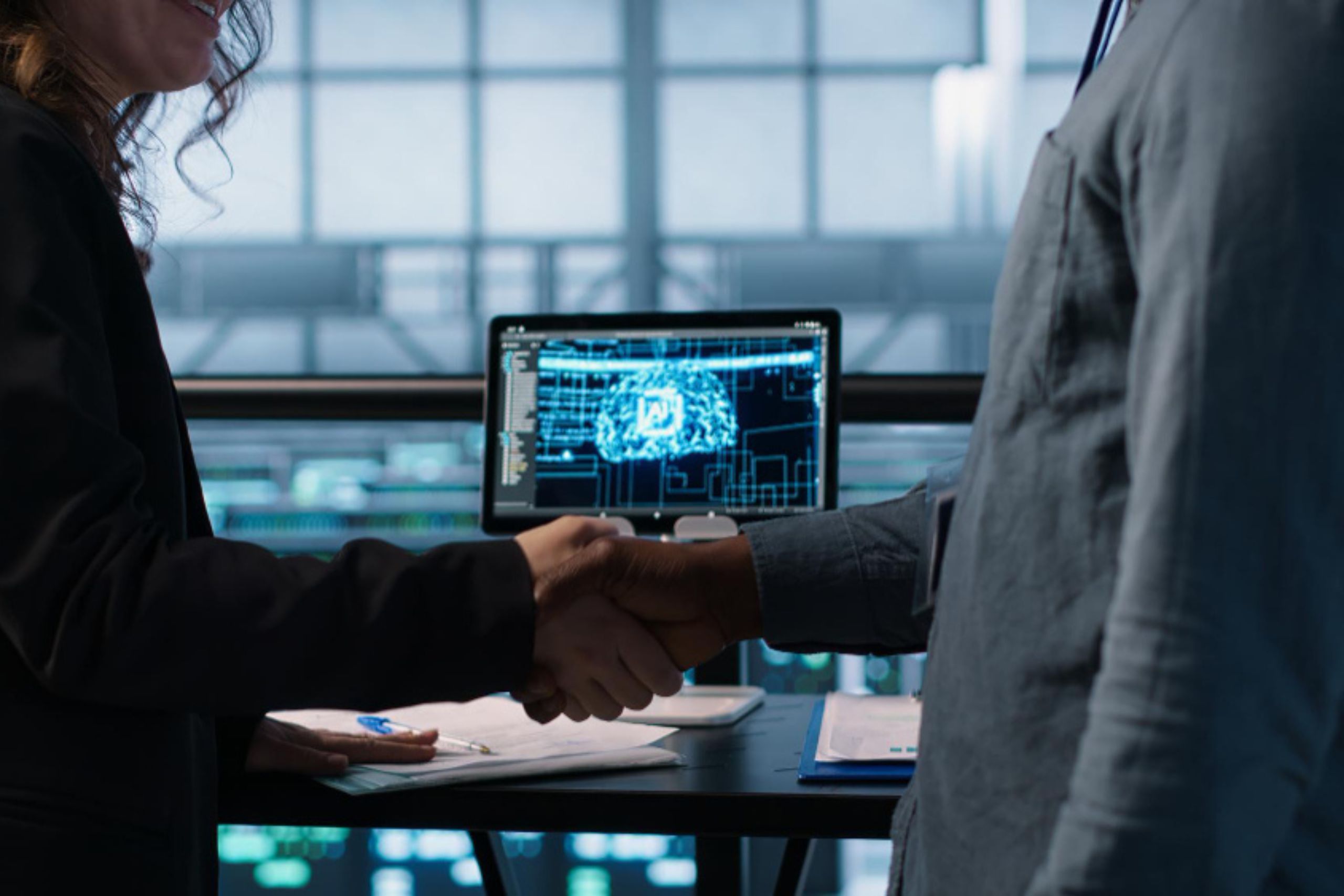 Two people shake hands in an office with a computer displaying a digital diagram in the background.