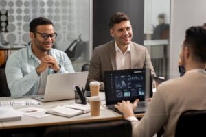 Three men sit at a table with laptops and coffee, discussing data displayed on a screen during a business meeting in a modern office.