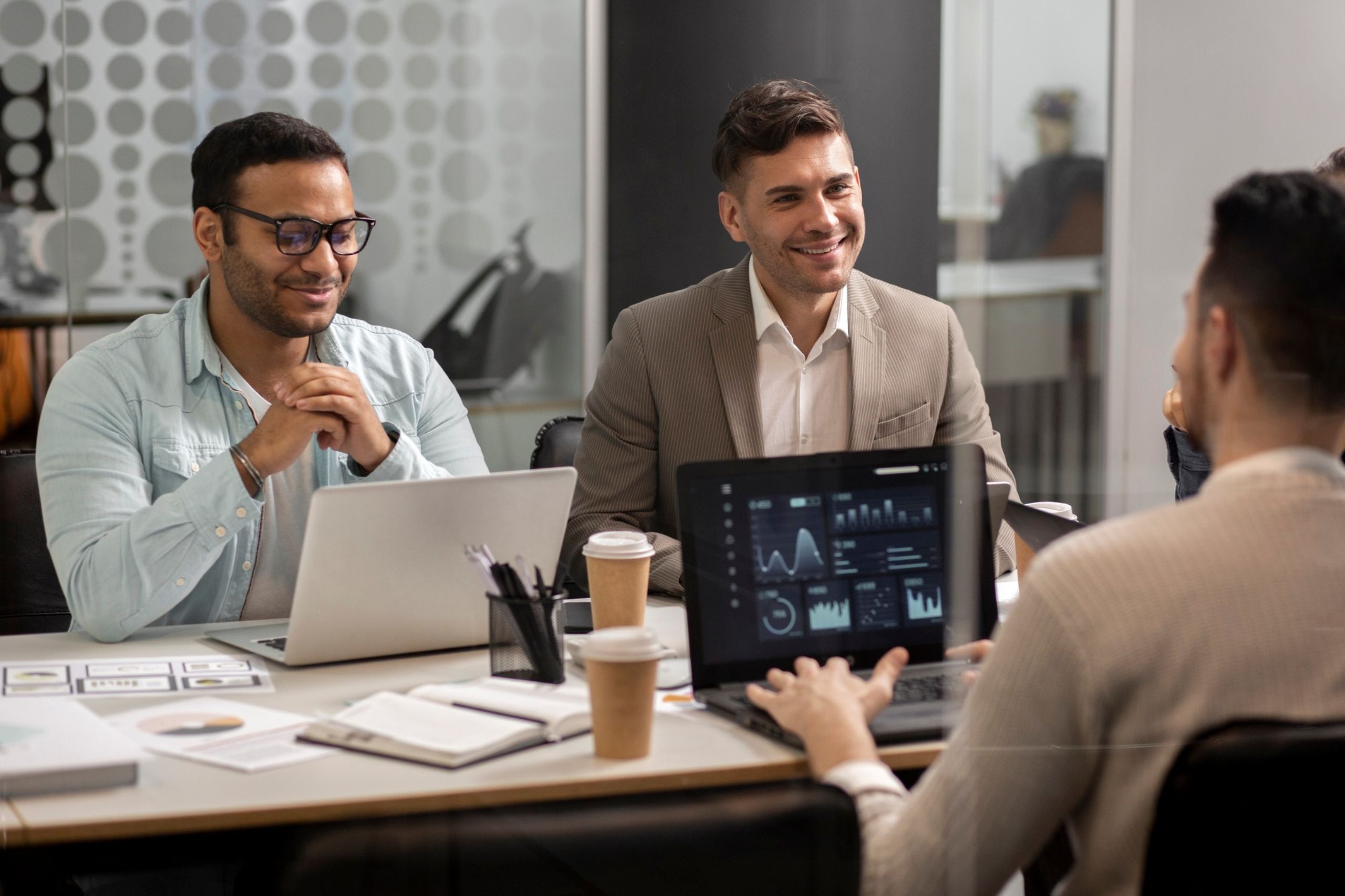 Three men sit at a table with laptops and coffee, discussing data displayed on a screen during a business meeting in a modern office.