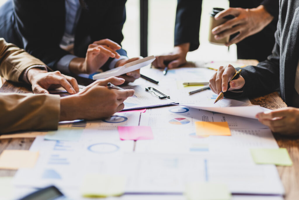 A group of people collaborates at a table covered with charts, graphs, documents, and colorful sticky notes, some using tablets and pens.