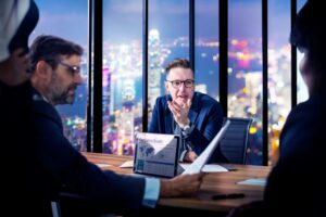 Four businesspeople in a high-rise office discuss company growth data, highlighting the importance of technology leadership for SMEs, with city lights visible through large windows in the background.
