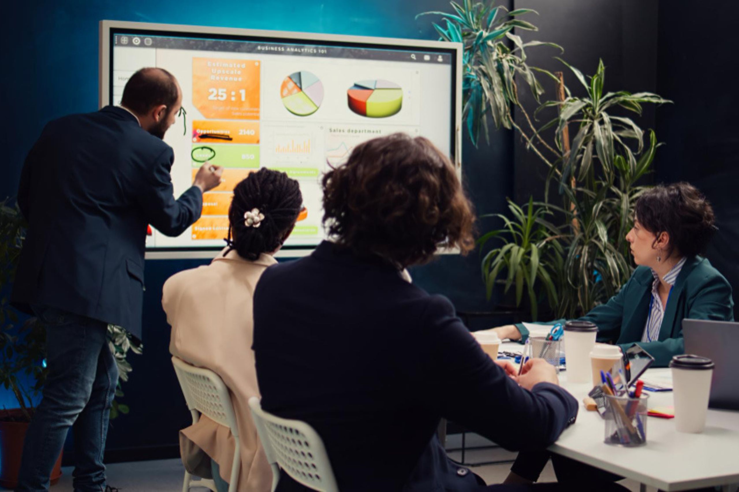A man presents charts and data on a large screen to three seated colleagues in a modern office with plants visible in the background, illustrating how an effective AI strategy for business growth can provide a significant competitive advantage.