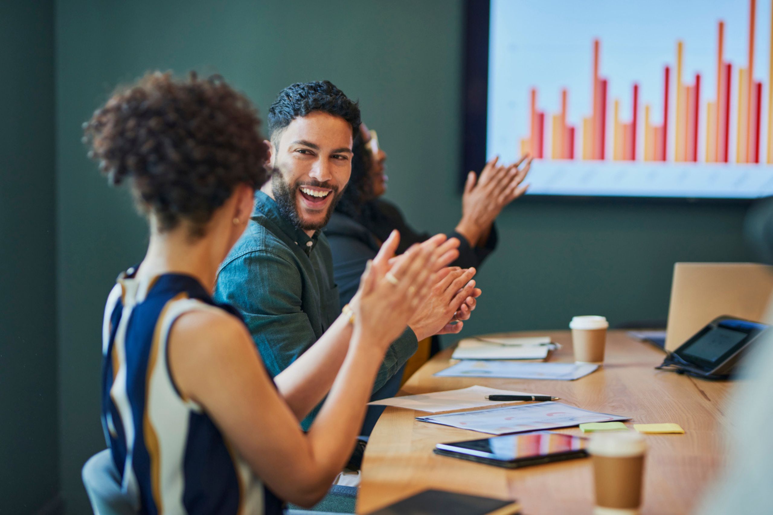 People seated at a conference table clapping, with a bar graph displayed on a screen in the background, celebrating the success of recent business performance solutions.