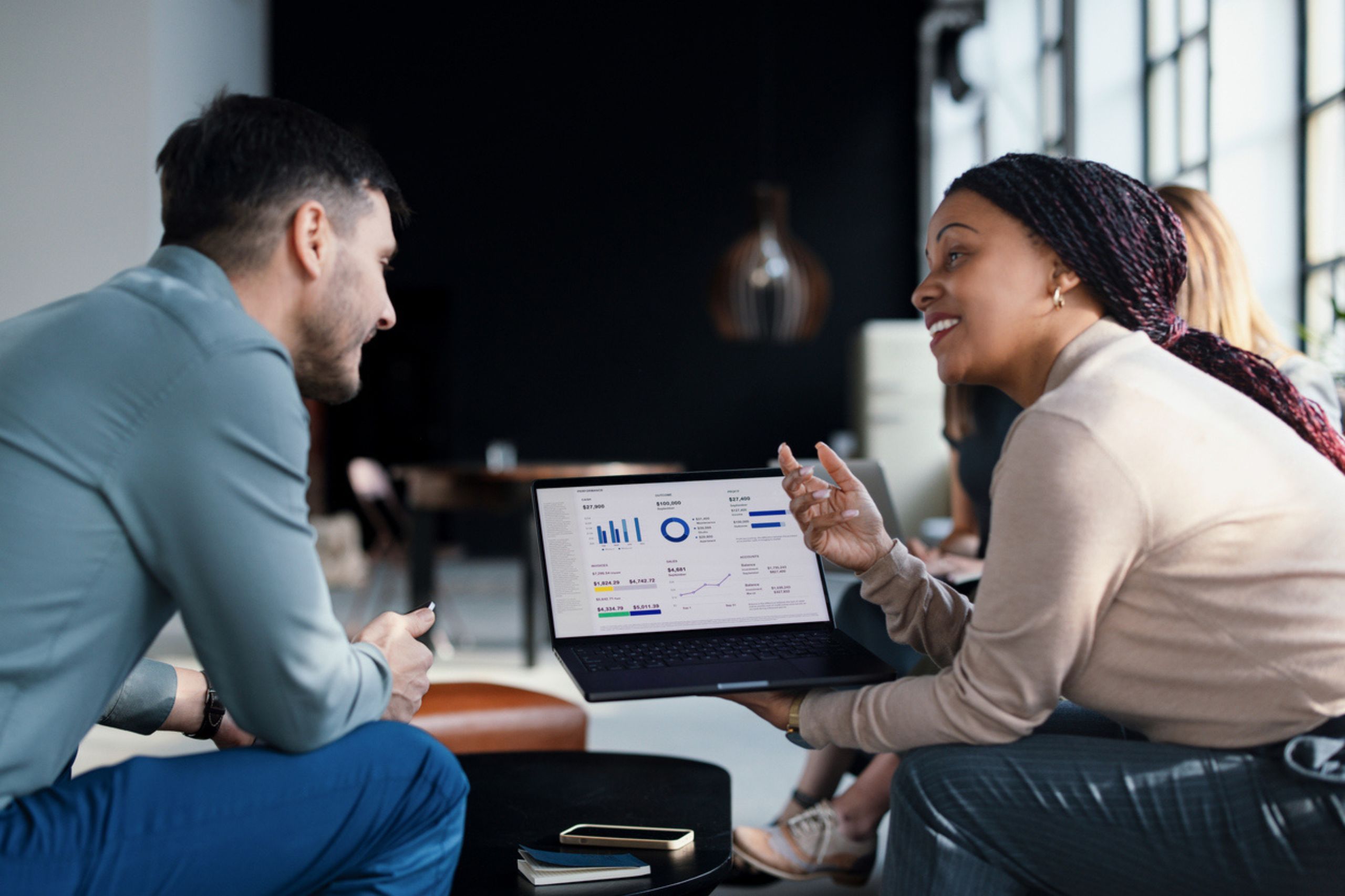 Two people sit indoors discussing business performance assessment as they analyze data displayed on a laptop screen showing graphs and charts, with notebooks and a phone on the table.