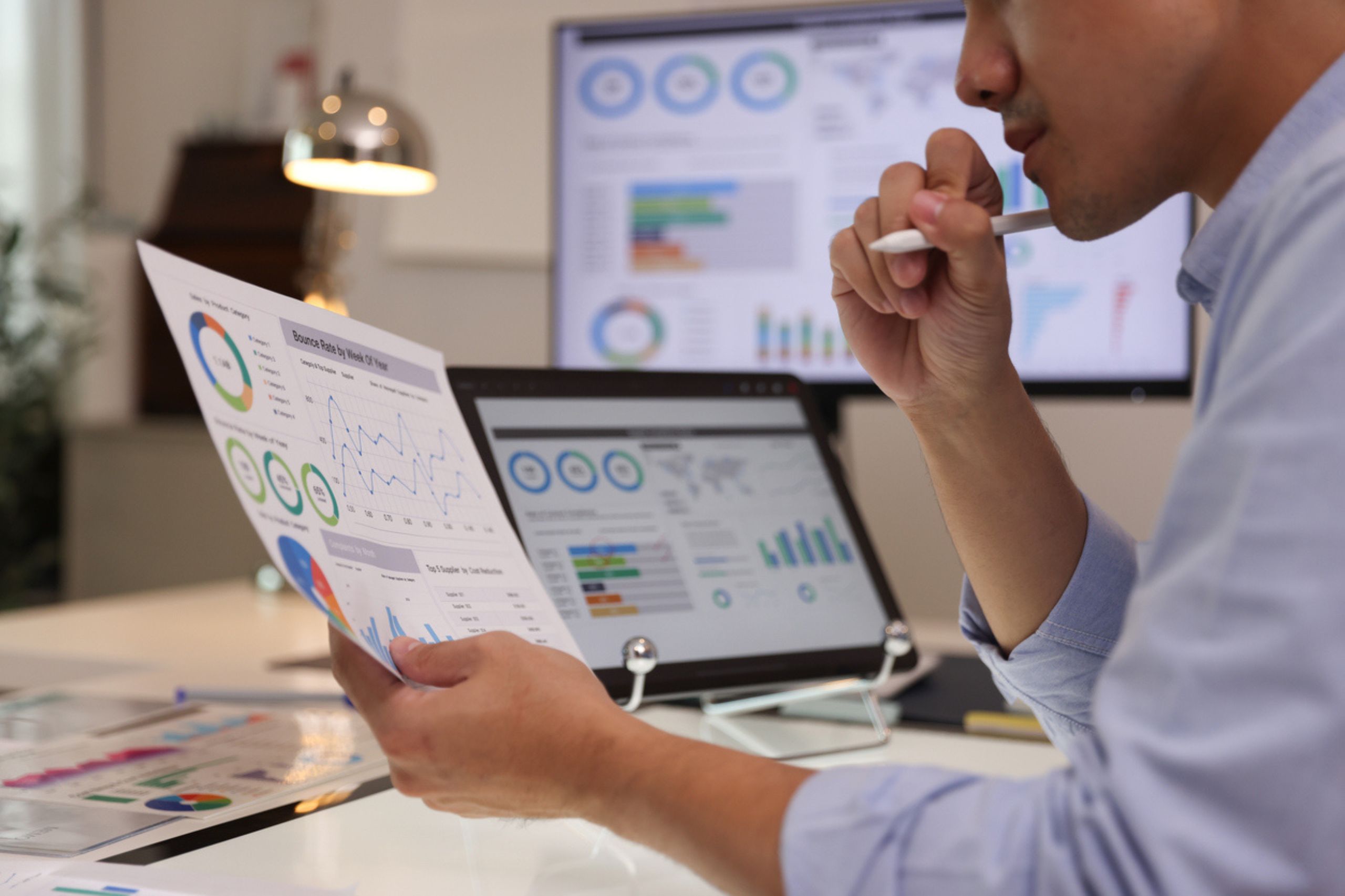 A person analyzes printed charts and graphs at a desk with a laptop and monitors displaying similar data in an office setting, focusing on operational efficiency to drive better business performance solutions.
