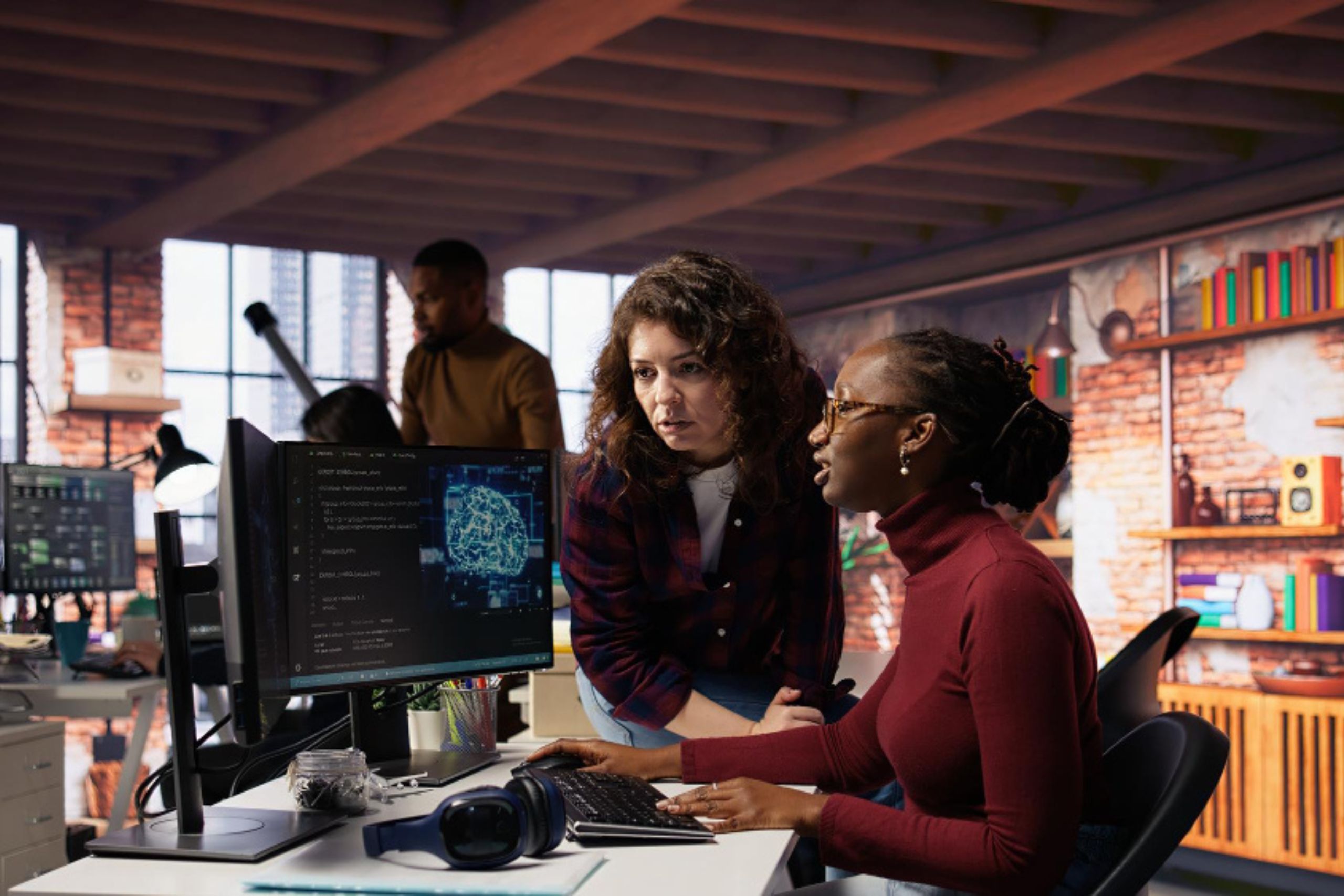 Two women in an office look at a computer screen displaying a digital brain image, while two other people work in the background.