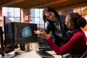 Two colleagues discuss a digital project at a desk with coding and a digital brain image on computer monitors in an office setting, collaborating on an AI readiness assessment as part of their organization's broader AI strategy.