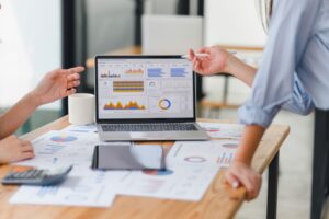 Two people discuss data shown on a laptop screen displaying charts and graphs, analyzing business performance assessments with documents, a tablet, a calculator, and a coffee mug on the wooden table.
