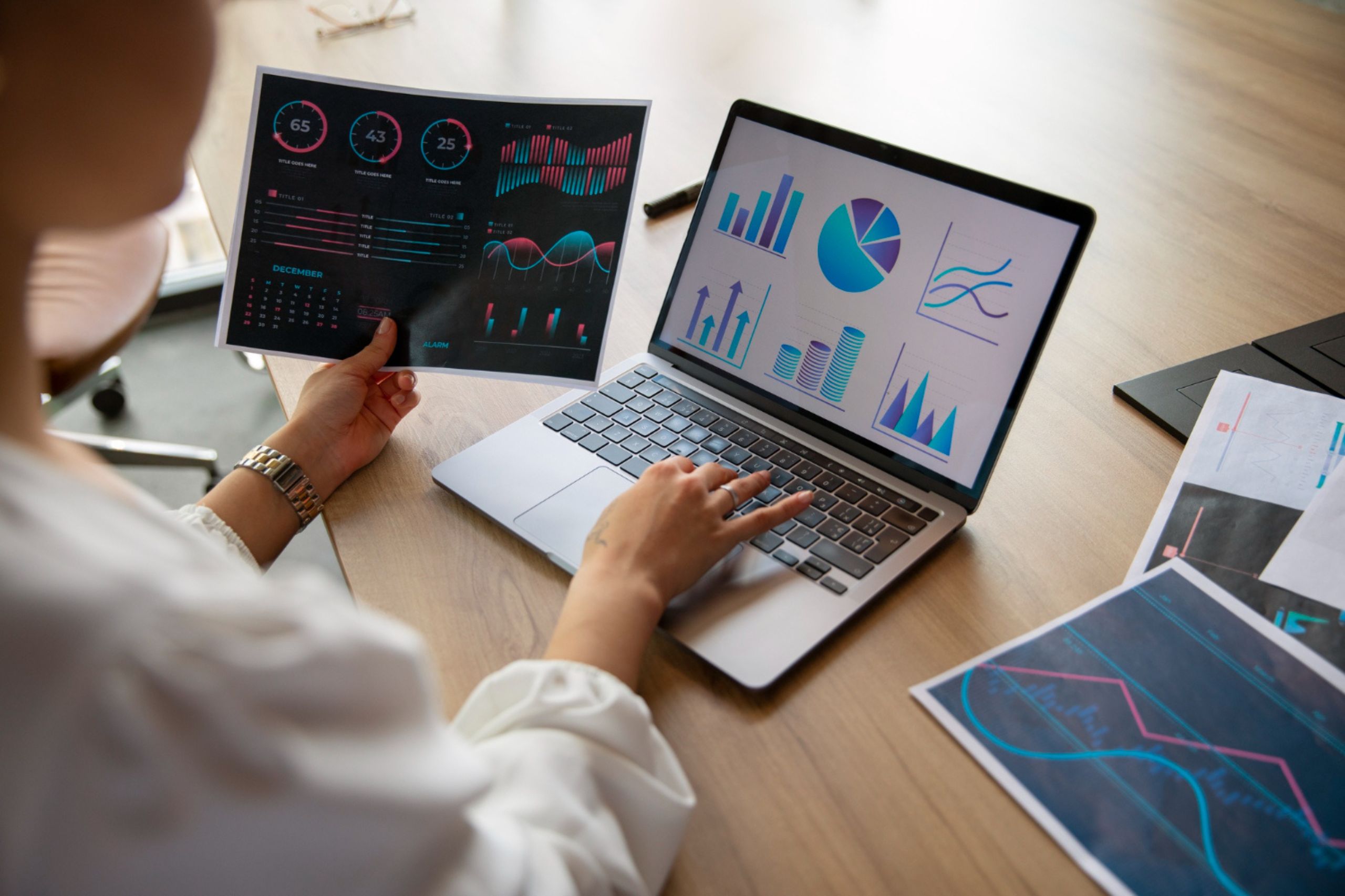 Person analyzing graphs and charts on a laptop and printed sheets at a desk, reviewing data visualizations and statistics to identify potential ai strategy mistakes and refine their ai implementation strategy.