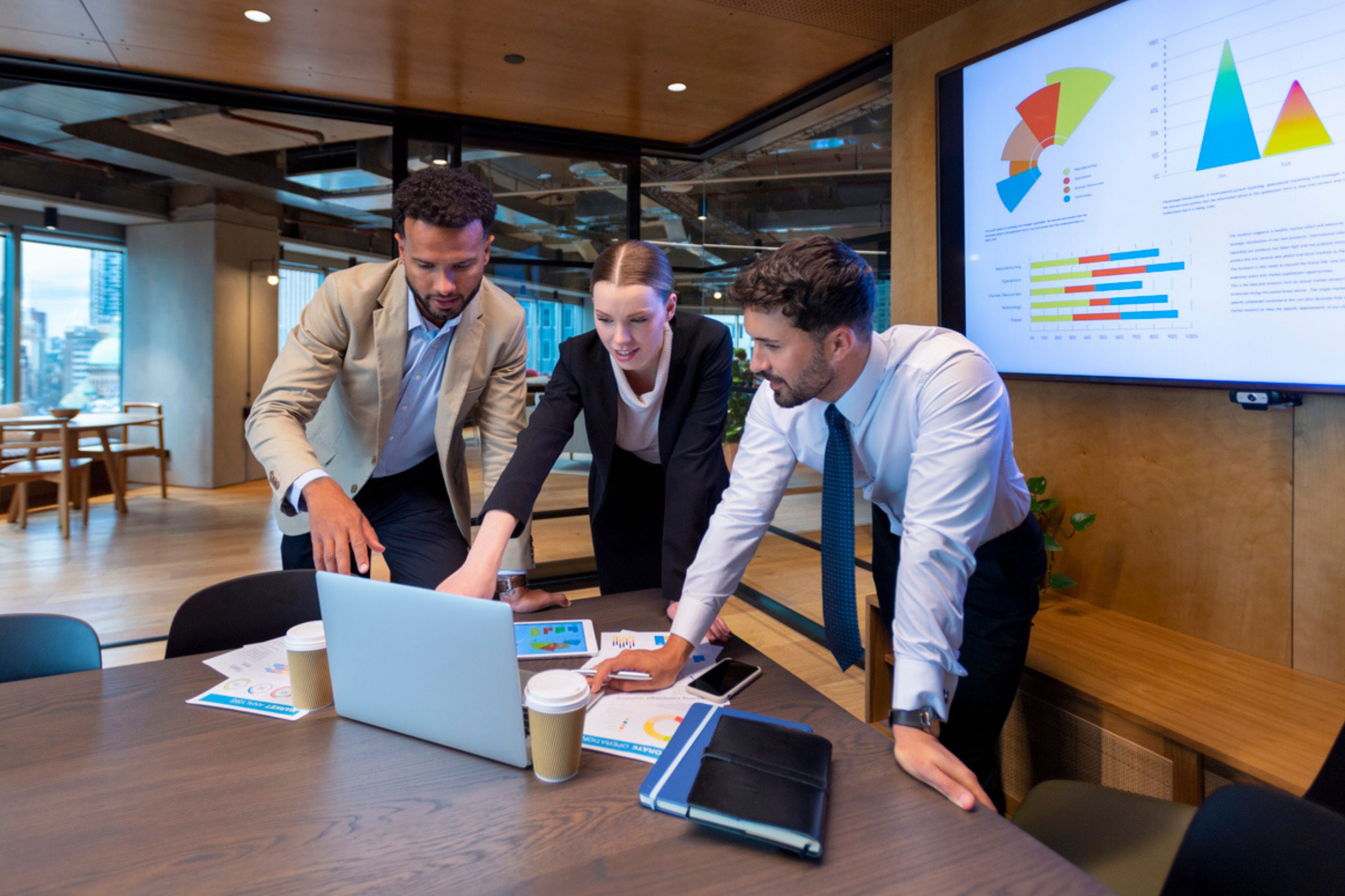 Three people stand around a table in a modern office, looking at a laptop while engaging in a business performance assessment. Charts and graphs are displayed on the wall screen behind them, illustrating key insights. Documents and coffee cups are scattered on the table as they discuss potential business performance solutions.