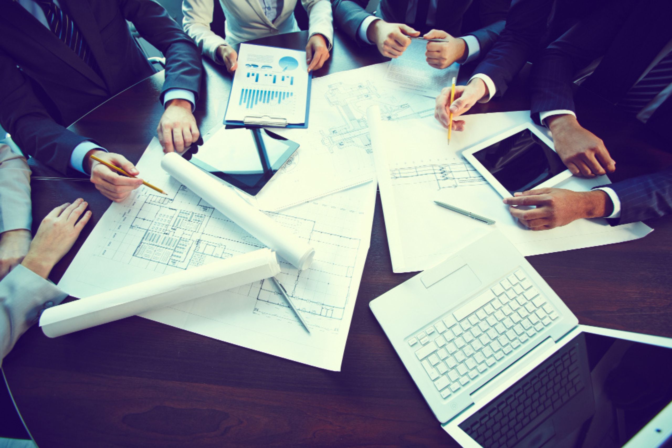 People in business attire discuss architectural blueprints at a table with laptops, tablets, and documents, collaboratively mapping out an ai roadmap to enhance their overall ai strategy.