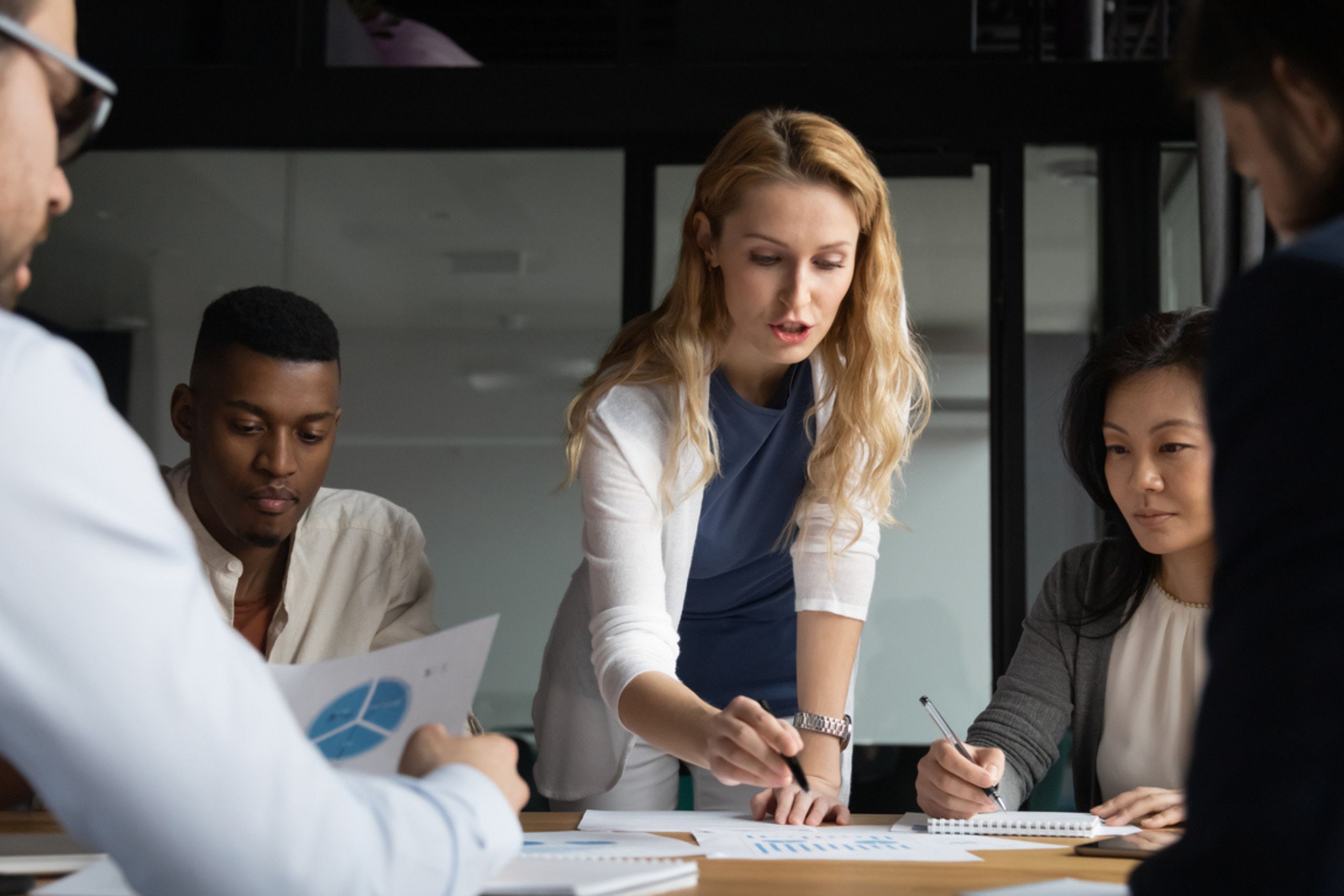 A woman stands and points to a paper on a table while three colleagues sit and listen during a business meeting focused on business performance assessment.