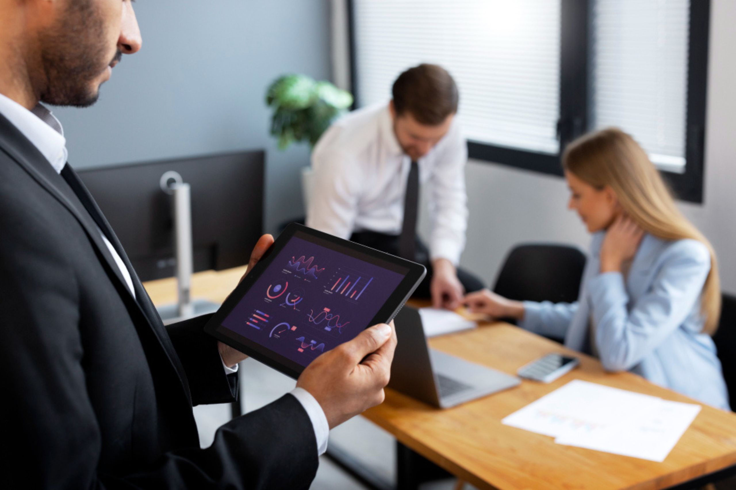 Man holds a tablet displaying charts from an AI readiness assessment, while two colleagues work together at a desk with papers and a laptop in a modern office.