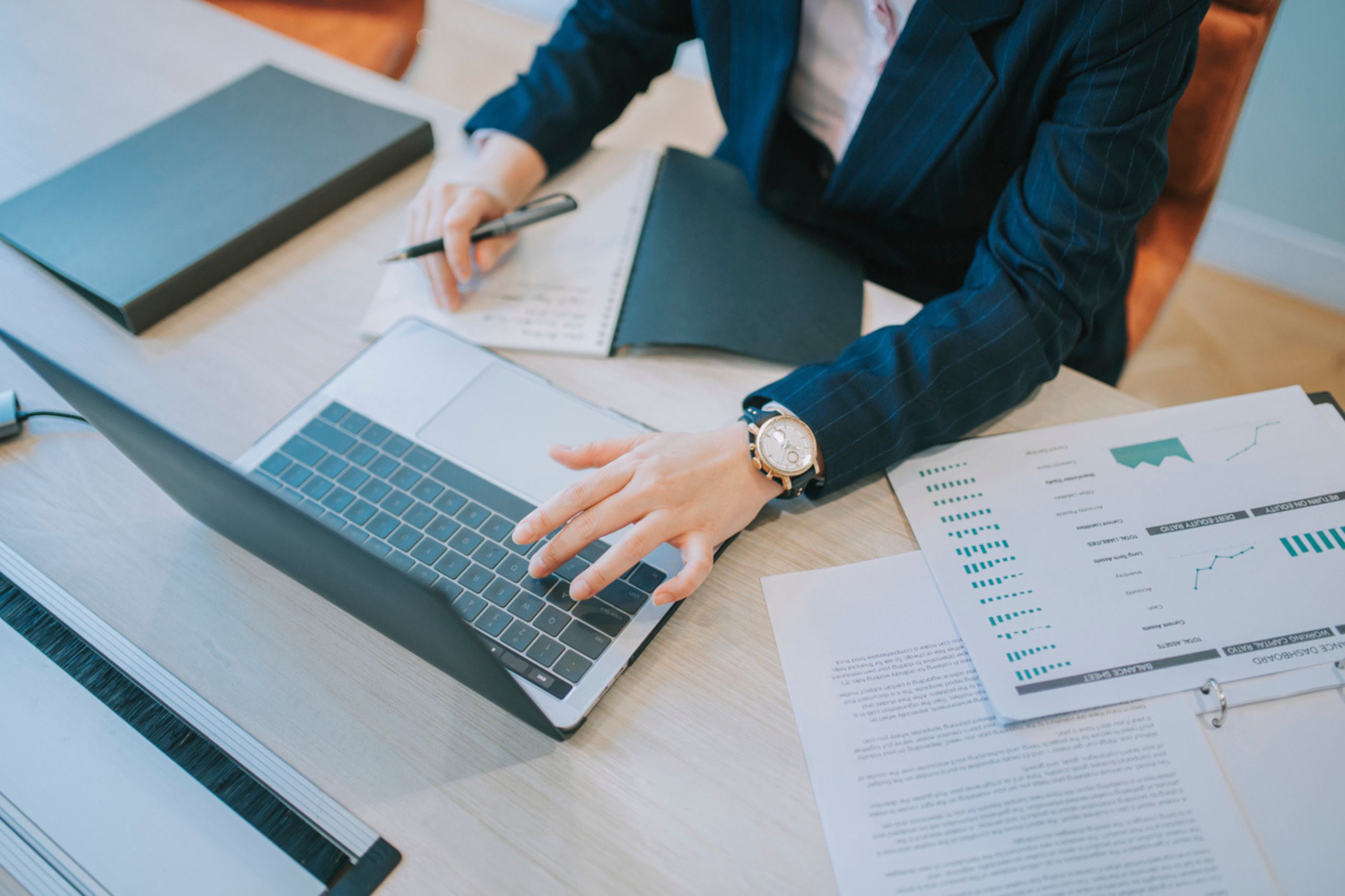 Person in business attire working at a desk with a laptop, notebook, and financial documents, writing notes and using the computer to measure business performance and explore Business Performance Solutions.