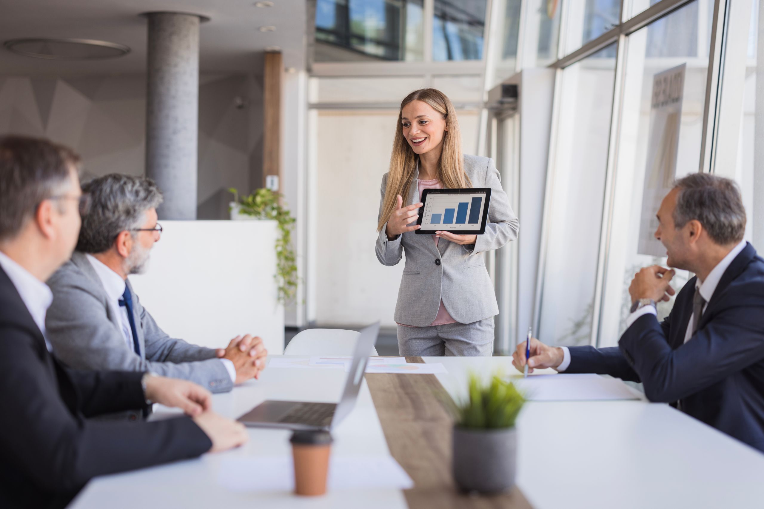 Woman in a suit presenting a tablet with a bar graph to three colleagues in a modern conference room, demonstrating effective business performance solutions.