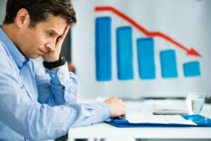 A man sits at a desk looking stressed, with his hand on his face. Behind him, a chart shows a downward trend with red arrow, illustrating business underperformance and highlighting the need for thorough business performance assessment.