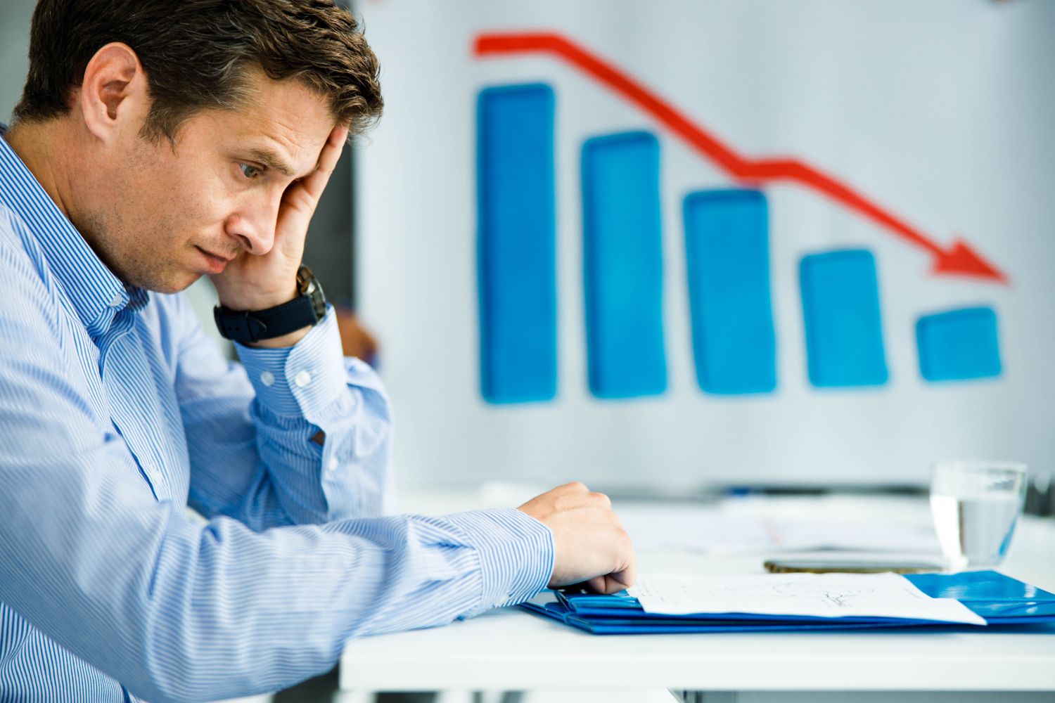 A man sits at a desk looking stressed, with his hand on his face. Behind him, a chart shows a downward trend with red arrow, illustrating business underperformance and highlighting the need for thorough business performance assessment.