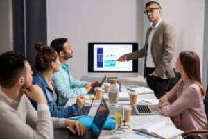 A man in a suit presents data on a screen to four colleagues seated at a table with laptops, notebooks, and coffee cups, illustrating the importance of technology leadership in driving digital transformation for SMEs.