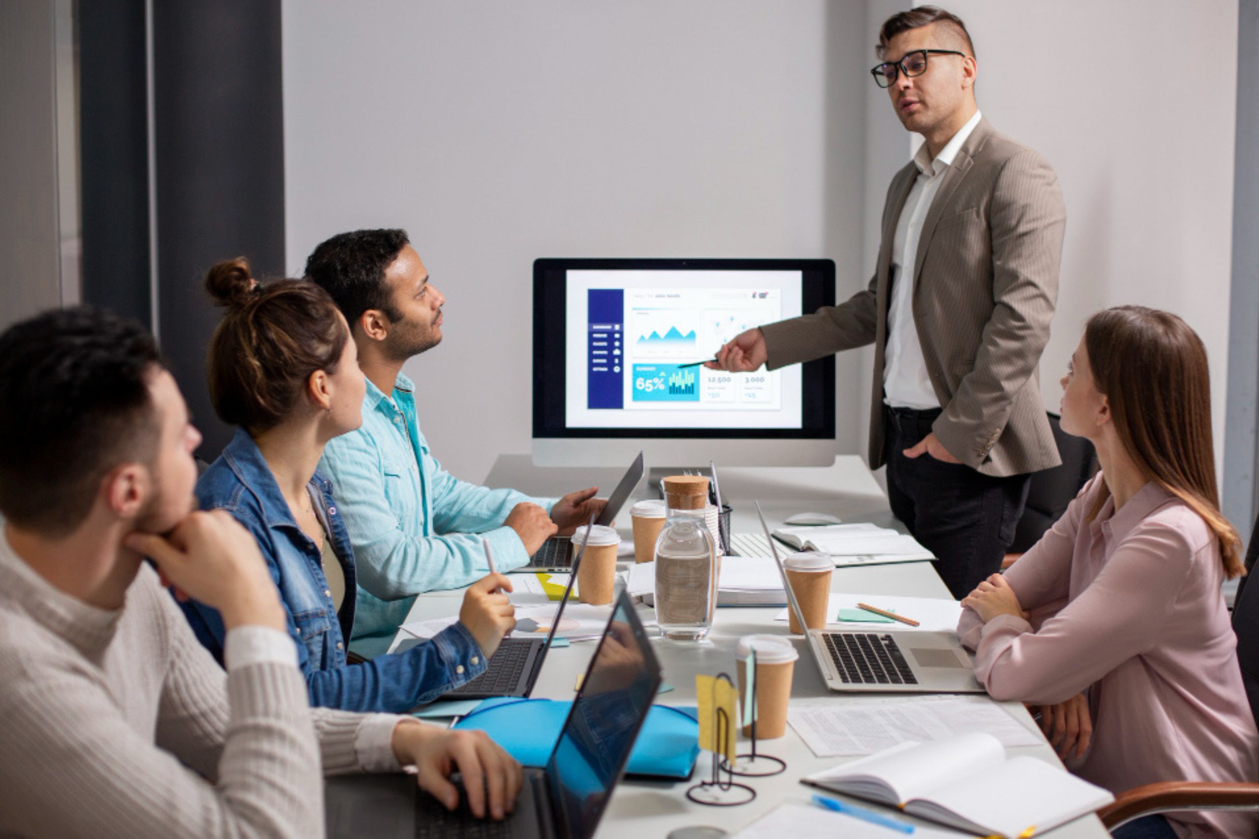 A man in a suit presents data on a screen to four colleagues seated at a table with laptops, notebooks, and coffee cups, illustrating the importance of technology leadership in driving digital transformation for SMEs.