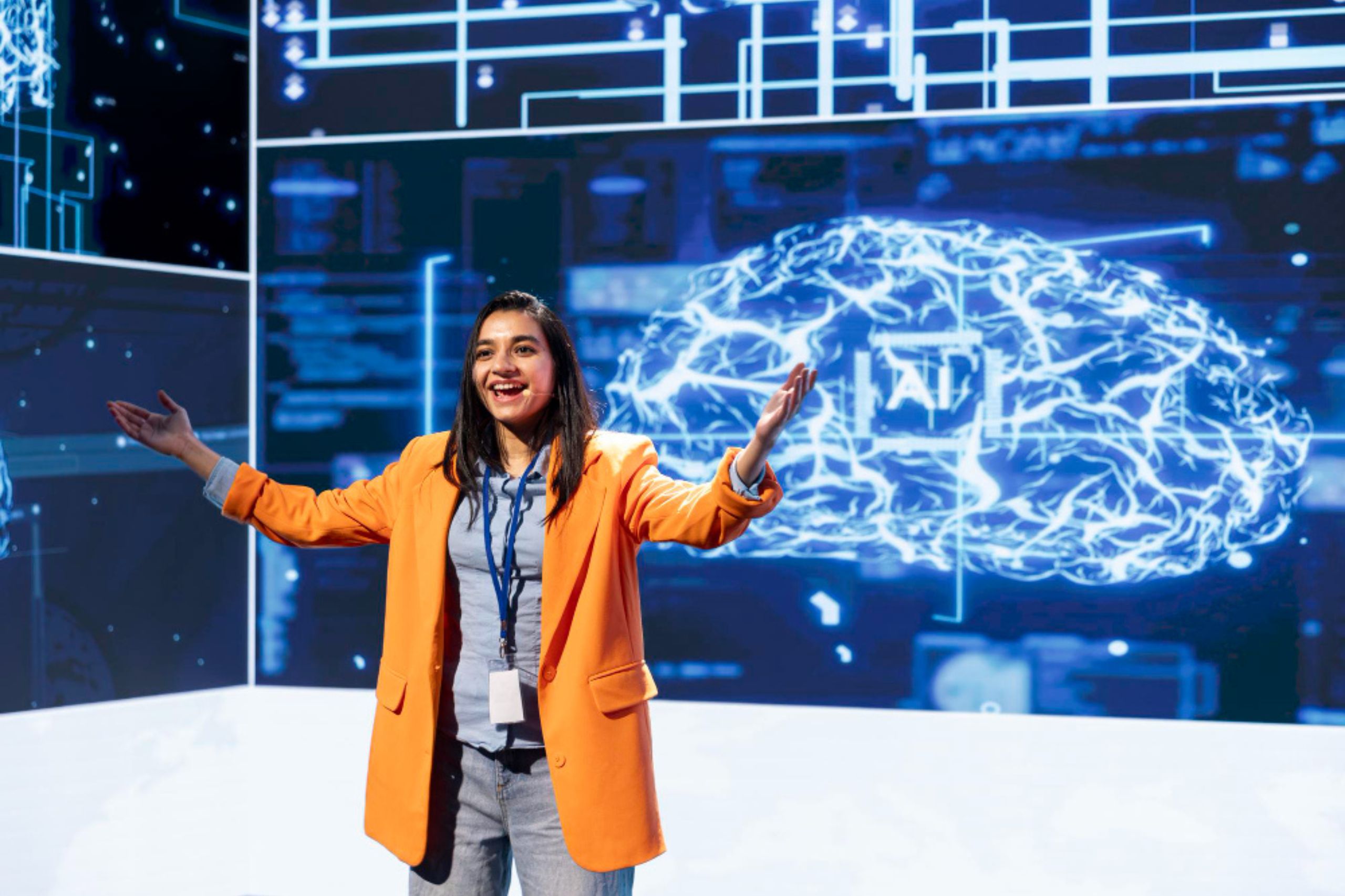 A woman in an orange blazer gestures while presenting on stage with a large digital brain graphic and AI-related visuals in the background, highlighting the importance of technology leadership & AI and advocating for responsible AI adoption.