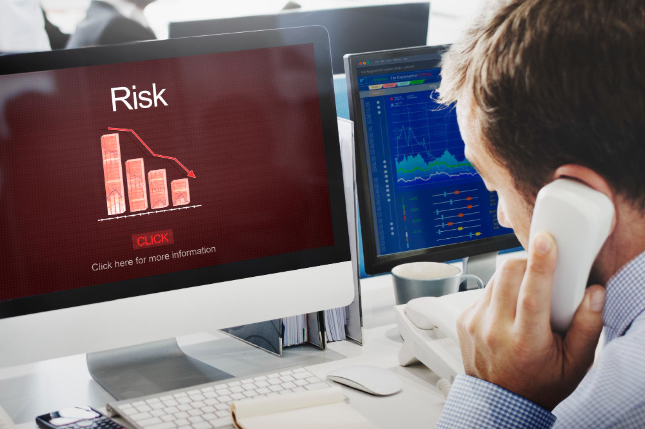 A man on the phone looks at a computer screen displaying a declining risk chart, with another monitor showing financial graphs beside him—an image that highlights technology leadership for SMEs as they navigate the challenges of digital transformation.