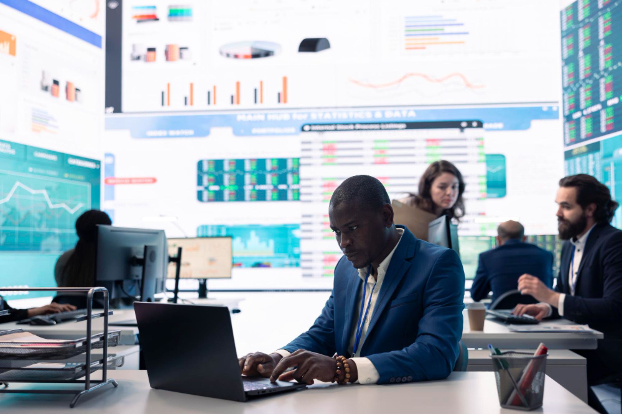 A man in a blue suit works on a laptop in an office with large digital screens displaying data, embodying technology leadership. Other professionals work in the background, highlighting the collaborative environment essential for driving digital transformation for SMEs.