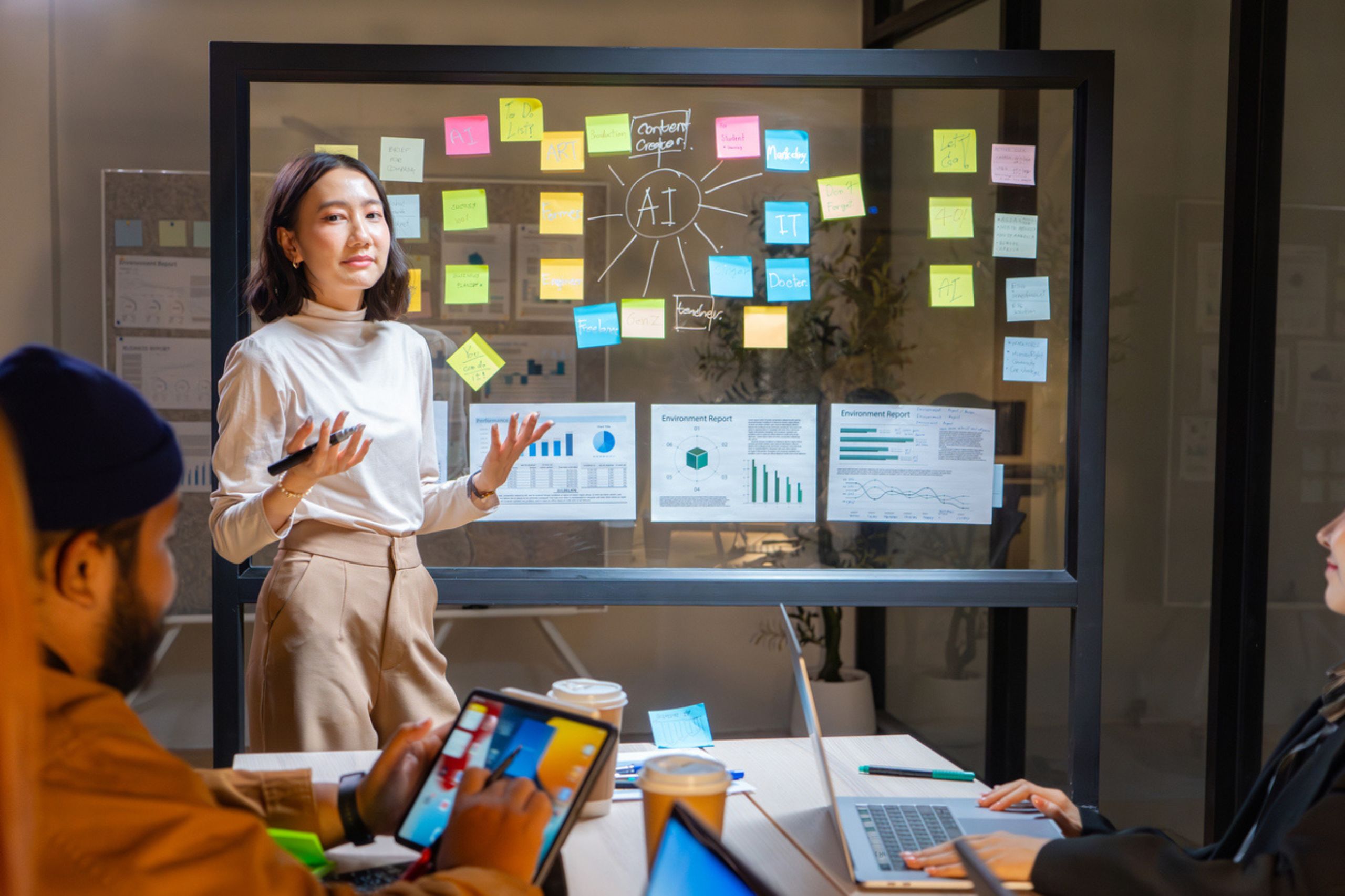 A woman gives a presentation to colleagues using a glass board with charts, graphs, and sticky notes, highlighting business performance solutions and digital transformation for SMEs, while others take notes on laptops at a table.