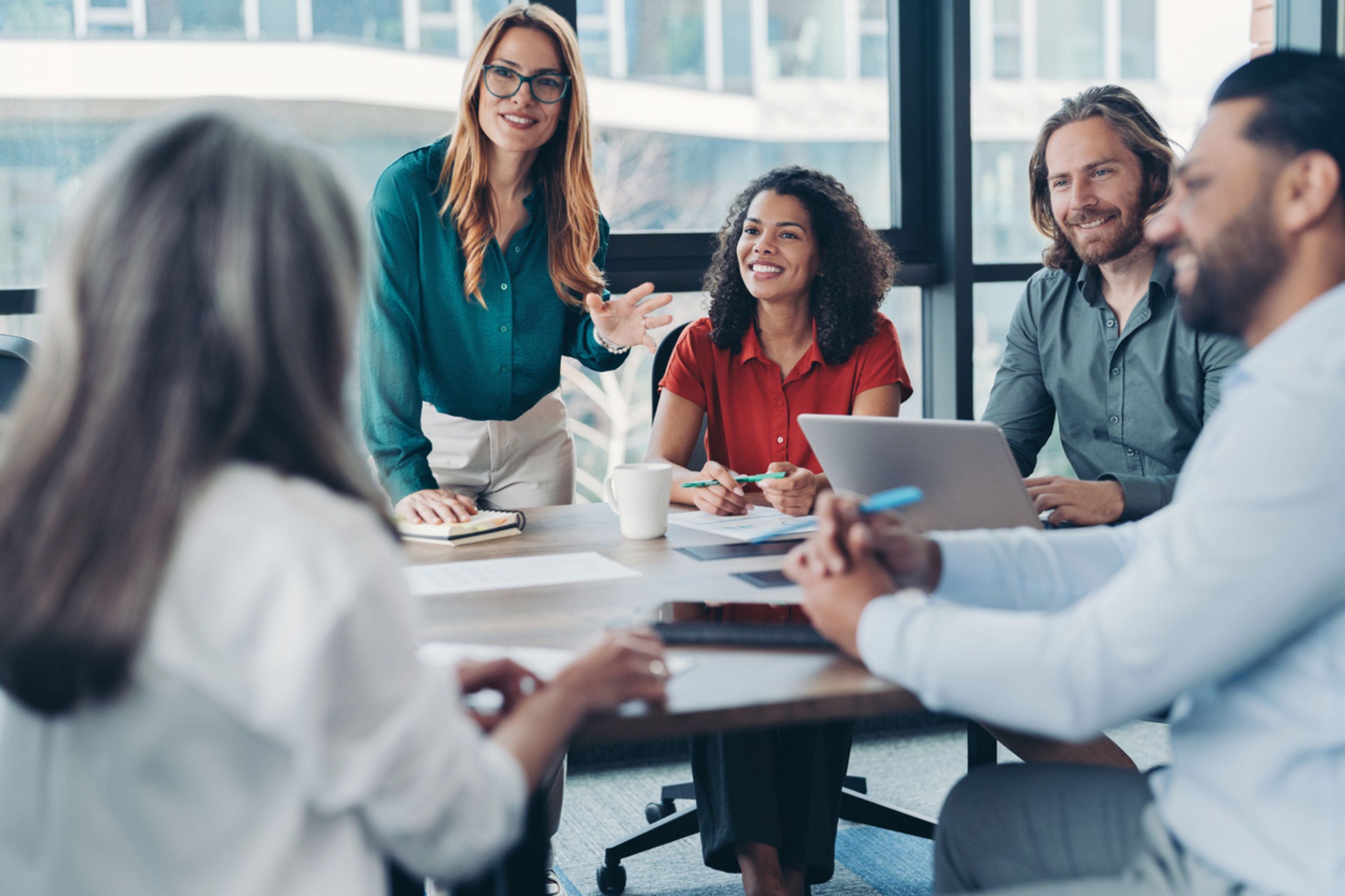 Five people sit around a table in a modern office, having a meeting focused on operational efficiency. One woman is standing and speaking, while the others are seated, listening and smiling. Laptops and papers related to business performance solutions are on the table.