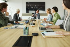 Five people in business attire sit around a conference table, engaging in a business performance assessment in front of a screen displaying a fluctuating line graph. Laptops, papers, and cups are on the table.