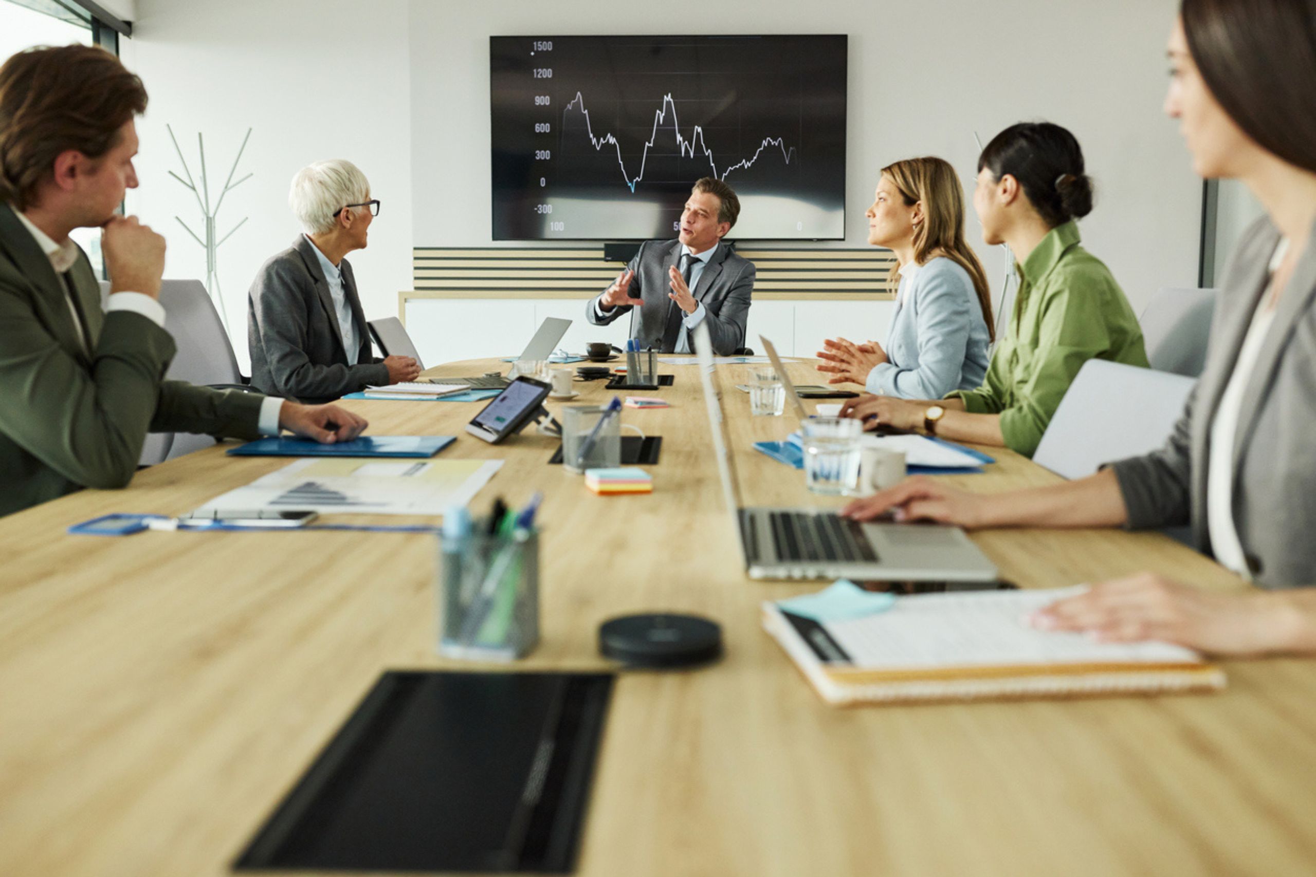 Five people in business attire sit around a conference table, engaging in a business performance assessment in front of a screen displaying a fluctuating line graph. Laptops, papers, and cups are on the table.