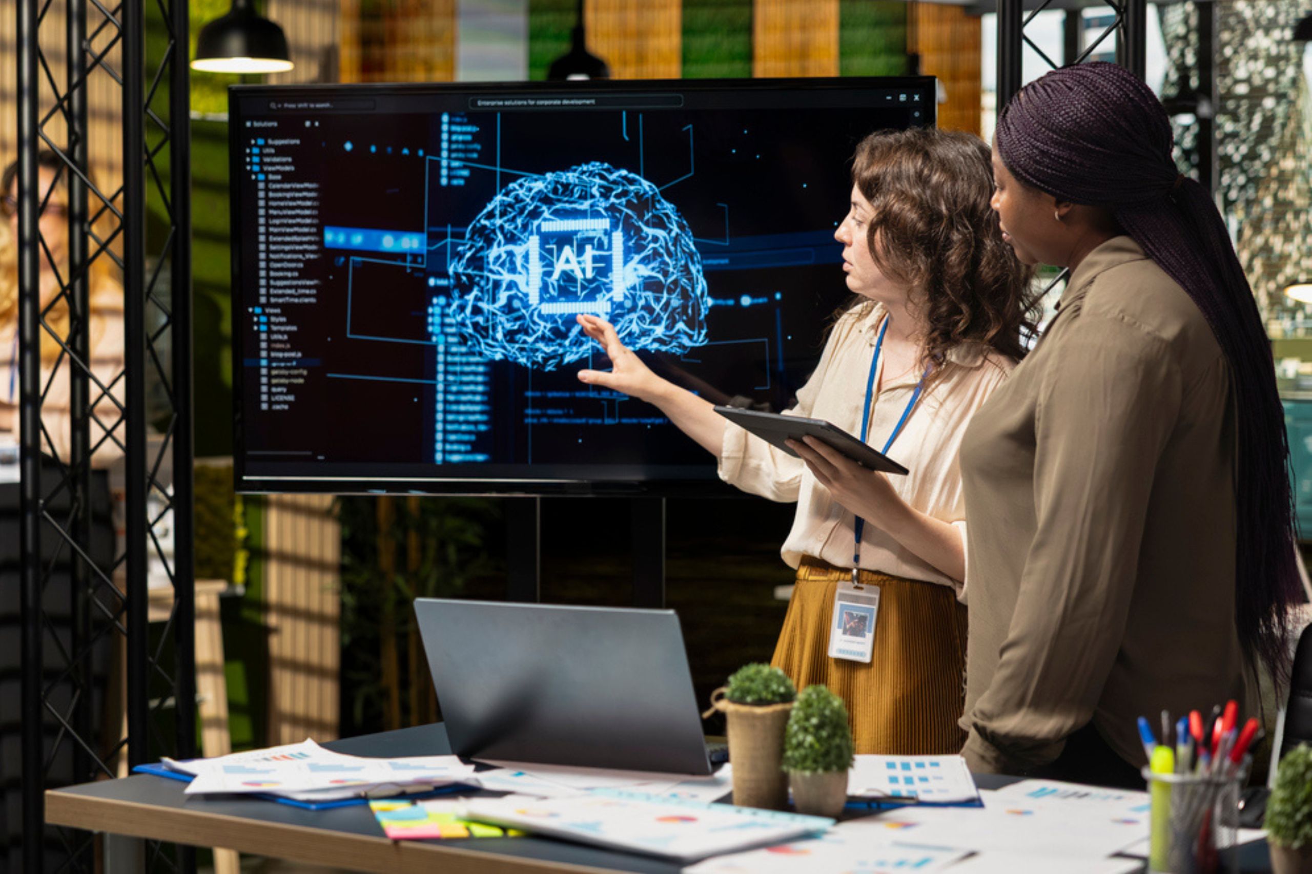 Two women stand by a desk, looking at a digital brain diagram on a large screen as they discuss their ai strategy. One points at the display while holding a tablet. Papers, a laptop, and plants are on the desk.