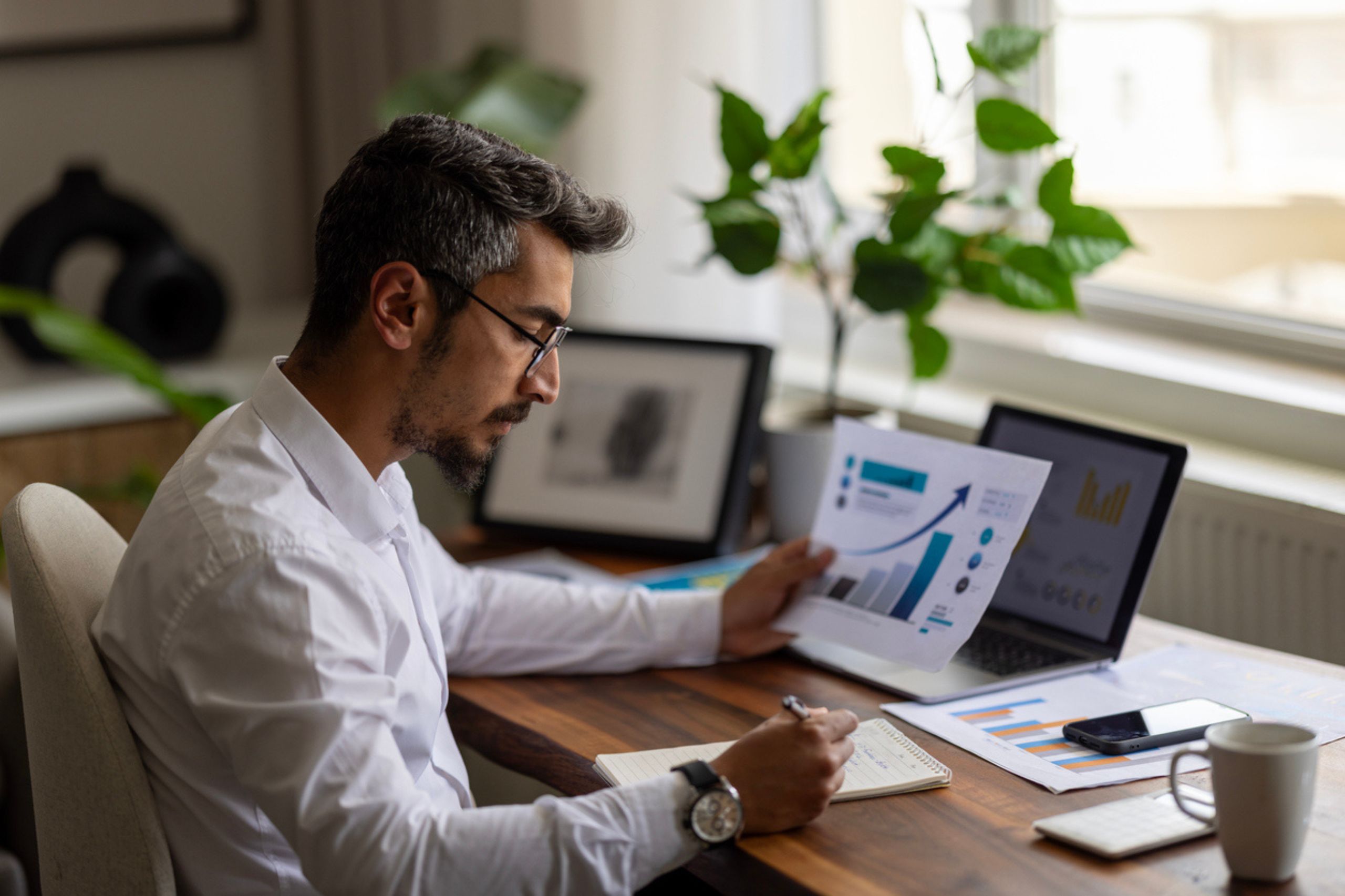 A man in a white shirt sits at a desk, reviewing financial charts and taking notes as he explores Business Performance Solutions, with a laptop, phone, and plants nearby.