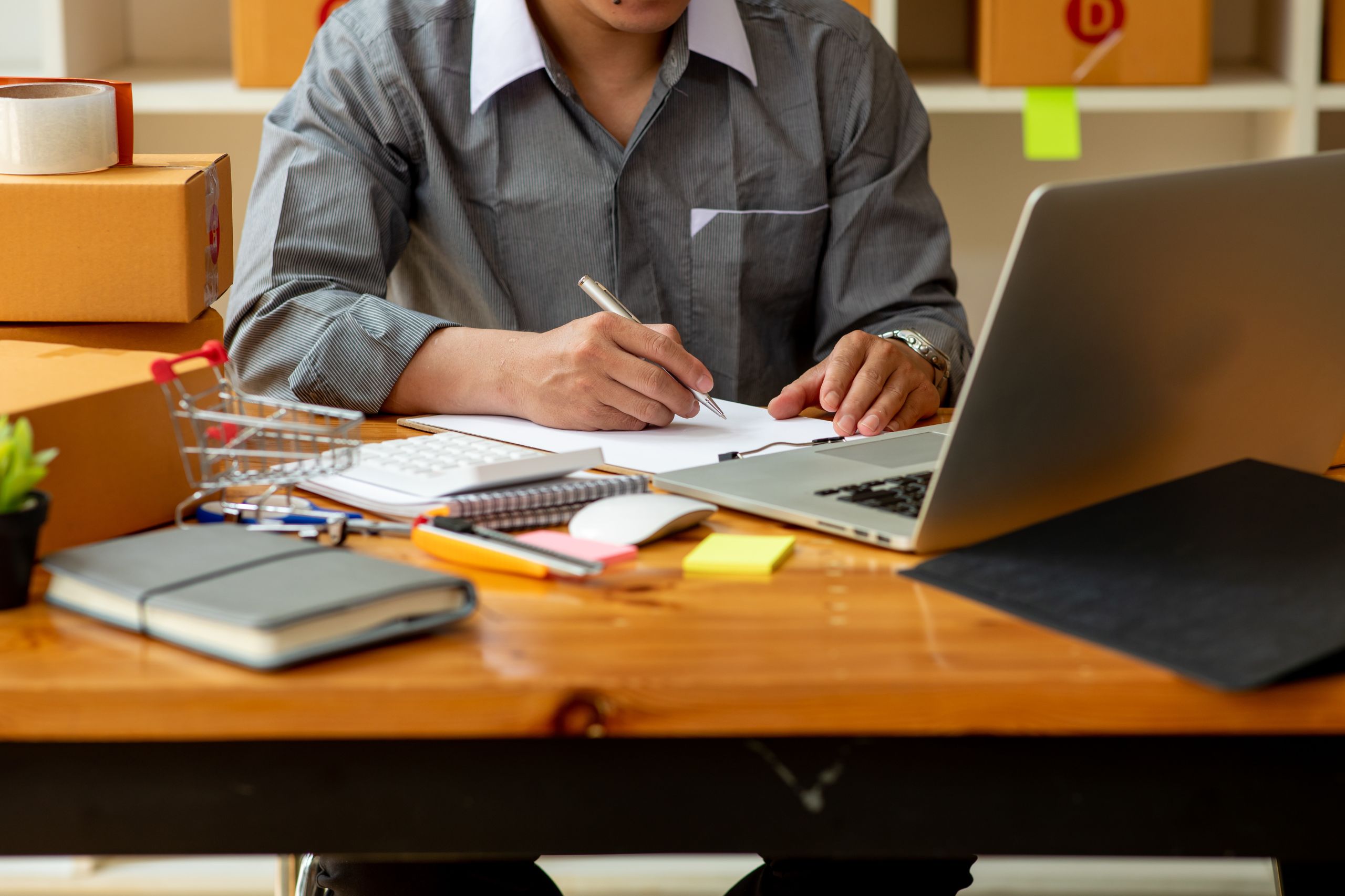 Man in a gray shirt writes on paper at a desk with a laptop, notebook, calculator, small shopping cart, and cardboard boxes—illustrating how Technology plays an essential role in streamlining business operations and supporting a successful Digital Transformation Strategy.