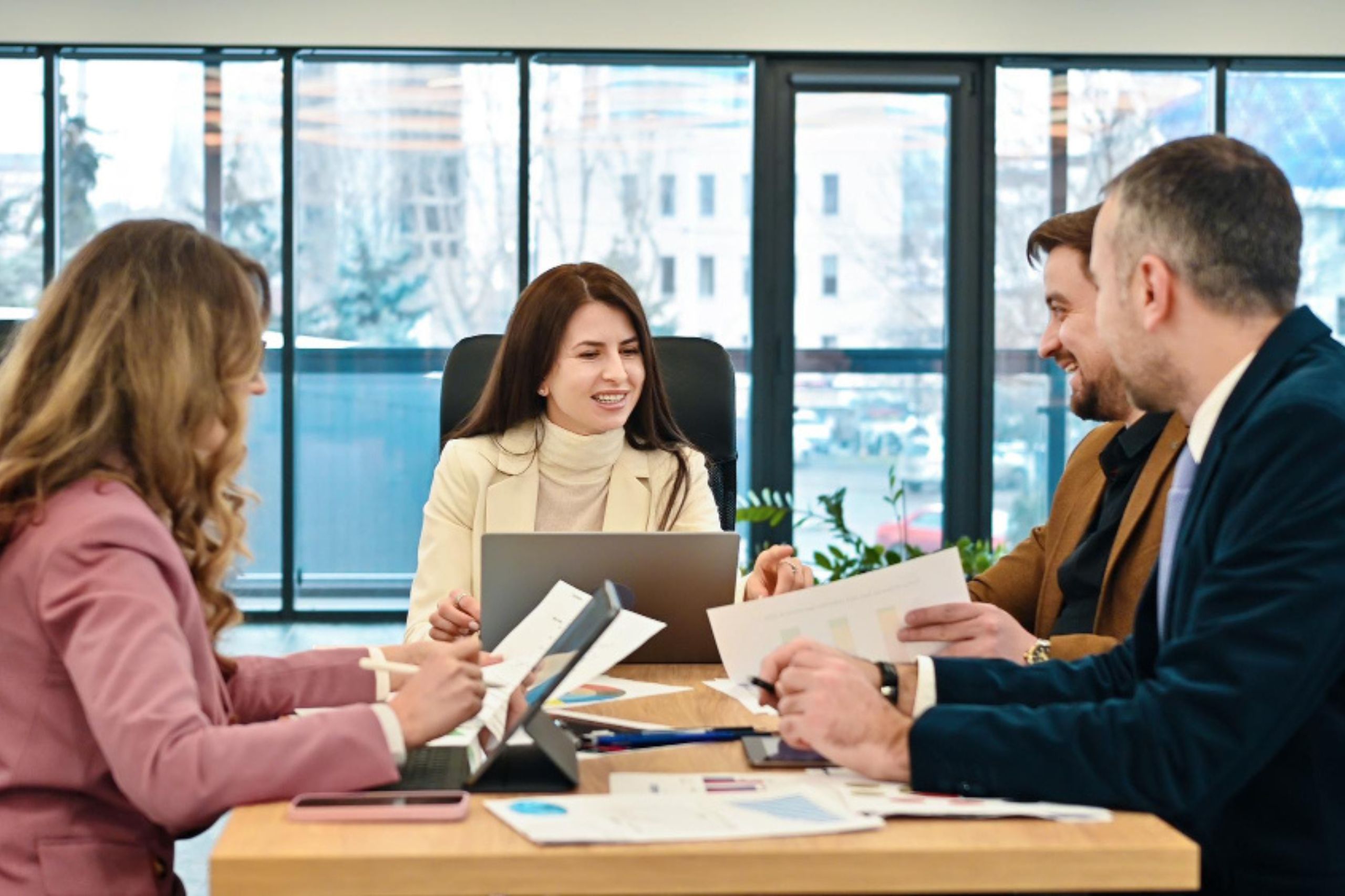 Four people in business attire sit around a table with laptops and papers, engaged in a meeting in a modern office with large windows in the background, discussing technology leadership for SMEs as they plan their next steps toward SME digital transformation.